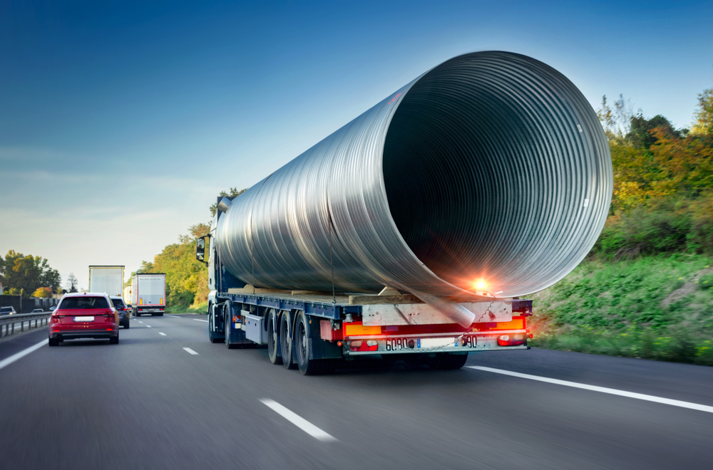 A highway truck carrying a massive cylindrical steel pipe with vehicles in the background.