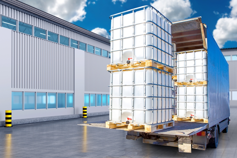 A freight truck loaded with large white industrial containers outside a warehouse.