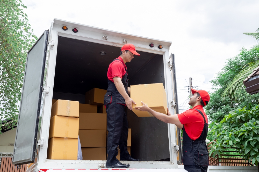 Two workers in red uniforms unloading cardboard boxes from a delivery truck outdoors.