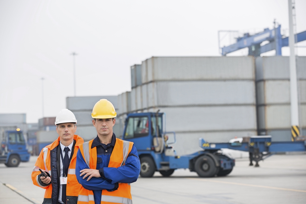 Two logistics professionals wearing safety vests and helmets standing in front of stacked shipping containers and industrial equipment.