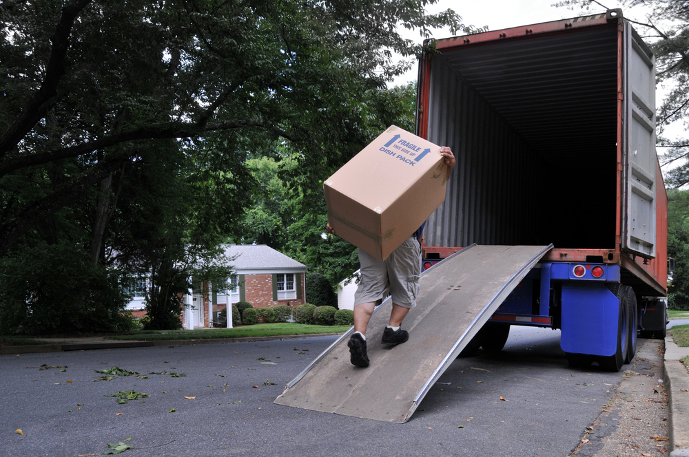 A worker carrying a large cardboard box up a ramp into a truck in a residential neighborhood.